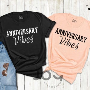 May include: Two t-shirts, one black and one peach, with the words "ANNIVERSARY Vibes" printed in white and black script. The black shirt is paired with blue jeans and sunglasses. The shirts are on a white wooden surface.