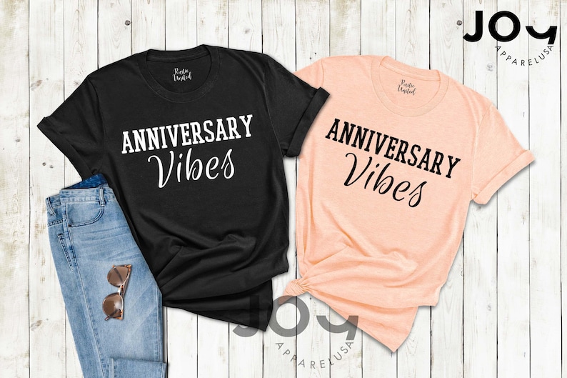 May include: Two t-shirts, one black and one peach, with the words "ANNIVERSARY Vibes" printed in white and black script. The black shirt is paired with blue jeans and sunglasses. The shirts are on a white wooden surface.