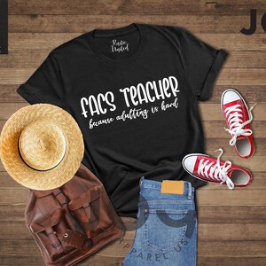May include: Black t-shirt with the text "FACS TEACHER because adulting is hard" in white. The image includes a straw hat, brown leather backpack, blue jeans, and red sneakers with white laces. The items are arranged on a wooden surface.