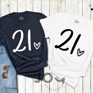 K&ouml;nnte beinhalten: Zwei T-Shirts, eines in Marineblau und eines in Wei&szlig;, jeweils mit der Zahl "21" und einem Herz. Das marineblaue Shirt hat wei&szlig;en Text, das wei&szlig;e Shirt schwarzen Text. Die Shirts werden mit einer Jeans und einer Sonnenbrille pr&auml;sentiert.