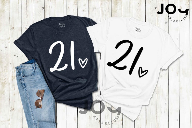 K&ouml;nnte beinhalten: Zwei T-Shirts, eines in Marineblau und eines in Wei&szlig;, jeweils mit der Zahl "21" und einem Herz. Das marineblaue Shirt hat wei&szlig;en Text, das wei&szlig;e Shirt schwarzen Text. Die Shirts werden mit einer Jeans und einer Sonnenbrille pr&auml;sentiert.