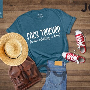 May include: A teal t-shirt with the text "FACS TEACHER because adulting is hard" is displayed with a straw hat, brown backpack, blue jeans, and red sneakers. The items are arranged on a wooden surface.