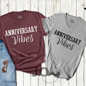 May include: Two t-shirts, one burgundy crew neck and one gray V-neck, both with the text "ANNIVERSARY Vibes" in white and black script. A pair of blue jeans and sunglasses are next to the burgundy shirt. The shirts are on a light wood background.