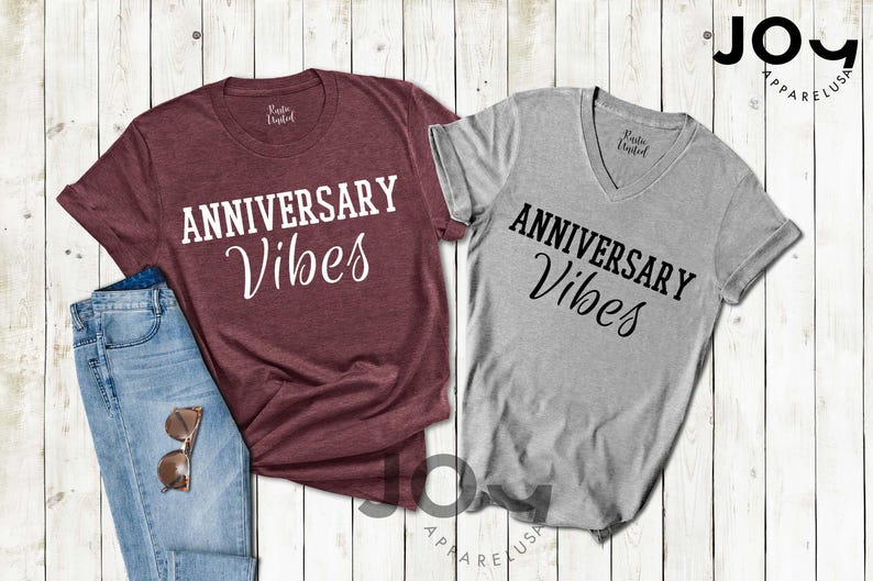 May include: Two t-shirts, one burgundy crew neck and one gray V-neck, both with the text "ANNIVERSARY Vibes" in white and black script. A pair of blue jeans and sunglasses are next to the burgundy shirt. The shirts are on a light wood background.
