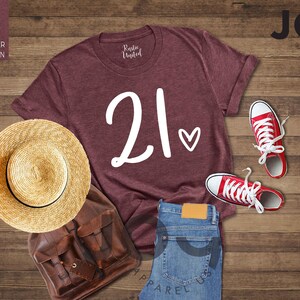 K&ouml;nnte beinhalten: Ein weinrotes T-Shirt mit der Zahl "21" und einem Herz, kombiniert mit roten Turnschuhen, einer blauen Jeans, einem Strohhut und einem braunen Rucksack. Das Shirt ist von "Rustic United" und "Joy Apparel US".