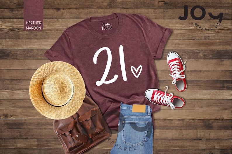 K&ouml;nnte beinhalten: Ein weinrotes T-Shirt mit der Zahl "21" und einem Herz, kombiniert mit roten Turnschuhen, einer blauen Jeans, einem Strohhut und einem braunen Rucksack. Das Shirt ist von "Rustic United" und "Joy Apparel US".