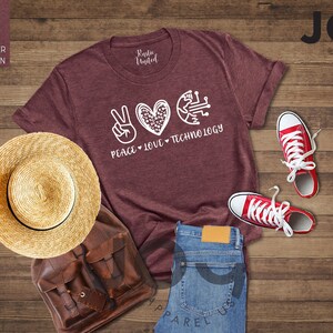 May include: A heather maroon t-shirt with the words "Peace, Love, Technology" and a peace sign, heart, and circuit board graphic. The image includes red sneakers, blue jeans, a straw hat, and a brown backpack.