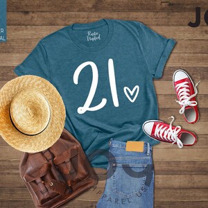 K&ouml;nnte beinhalten: Ein tealfarbenes T-Shirt mit der Zahl "21" und einem Herz-Motiv, kombiniert mit roten Turnschuhen, einem Strohhut, einem braunen Rucksack und einer blauen Jeans. Die Artikel sind auf einer Holzoberfl&auml;che angeordnet.