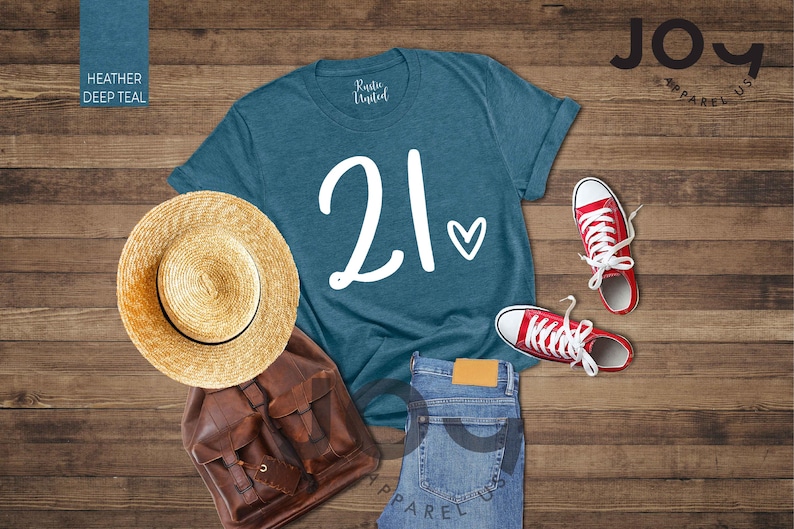 K&ouml;nnte beinhalten: Ein tealfarbenes T-Shirt mit der Zahl "21" und einem Herz-Motiv, kombiniert mit roten Turnschuhen, einem Strohhut, einem braunen Rucksack und einer blauen Jeans. Die Artikel sind auf einer Holzoberfl&auml;che angeordnet.