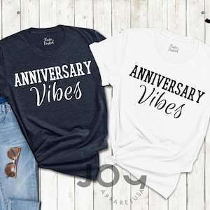 May include: Two t-shirts, one navy and one white, with the words "ANNIVERSARY Vibes" printed in black. The navy shirt is paired with blue jeans and sunglasses. The shirts are from Joy Apparel USA.