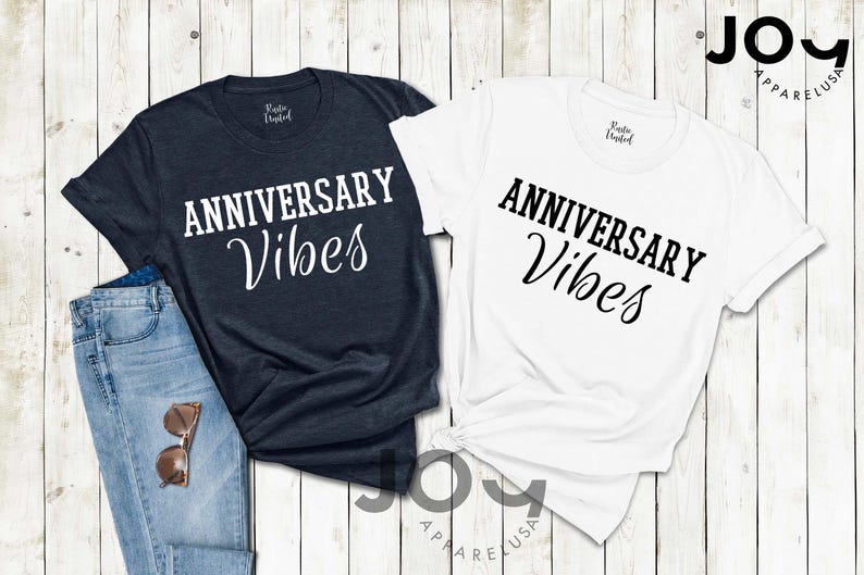 May include: Two t-shirts, one navy and one white, with the words "ANNIVERSARY Vibes" printed in black. The navy shirt is paired with blue jeans and sunglasses. The shirts are from Joy Apparel USA.