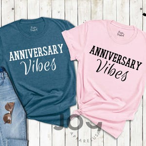 May include: Two t-shirts, one teal and one pink, with the words "ANNIVERSARY Vibes" printed in white and black, respectively. The teal shirt is paired with blue jeans and sunglasses. The shirts are from Joy Apparel USA.