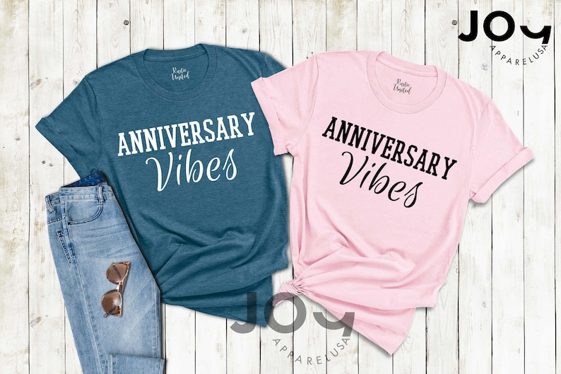 May include: Two t-shirts, one teal and one pink, with the words "ANNIVERSARY Vibes" printed in white and black, respectively. The teal shirt is paired with blue jeans and sunglasses. The shirts are from Joy Apparel USA.