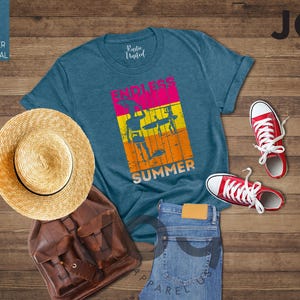 May include: A teal t-shirt with "ENDLESS SUMMER" text and graphic, straw hat, brown backpack, blue jeans, and red sneakers on a wood background. The shirt has a retro design with surfers. A casual summer outfit.