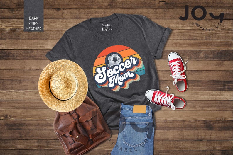 May include: Dark grey heather t-shirt with a retro "Soccer Mom" graphic, paired with red trainers, blue jeans, a straw hat, and a brown backpack. The shirt features a soccer ball design with a colorful sunset background.