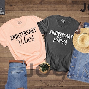 May include: Two t-shirts, one heather peach and one dark grey heather, with "ANNIVERSARY Vibes" printed in black script. A denim jacket, straw hat, and sunglasses are also displayed. A pair of blue jeans with a brown belt and a small potted plant complete the arrangement.