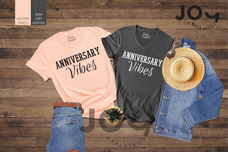 May include: Two t-shirts, one heather peach and one dark grey heather, with "ANNIVERSARY Vibes" printed in black script. A denim jacket, straw hat, and sunglasses are also displayed. A pair of blue jeans with a brown belt and a small potted plant complete the arrangement.
