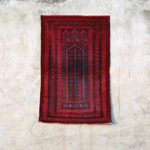 May include: A rectangular, red and black rug with an intricate geometric pattern. The rug features a central design with a dark blue background and a red border. The rug is displayed against a textured, off-white wall.