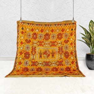May include: A rectangular, golden-yellow rug with a detailed, repeating pattern of red and cream-colored floral and geometric designs. The rug features a central rectangular panel with figures. The rug is hanging against a white wall, with a potted plant to the right.