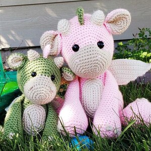 May include: Two crocheted stuffed animals, one pink and one green, sitting in grass. The pink animal has wings and a horn, while the green animal has a horn and a tail.