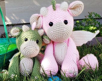May include: Two crocheted stuffed animals, one pink and one green, sitting in grass. The pink animal has wings and a horn, while the green animal has a horn and a tail.