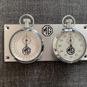 Może przedstawiać: Dwa srebrne stopery zamontowane na płycie ze szczotkowanego metalu. Stoperki mają białe tarcze z czarnymi i czerwonymi oznaczeniami oraz logo MG. Płyta ma logo MG pośrodku.