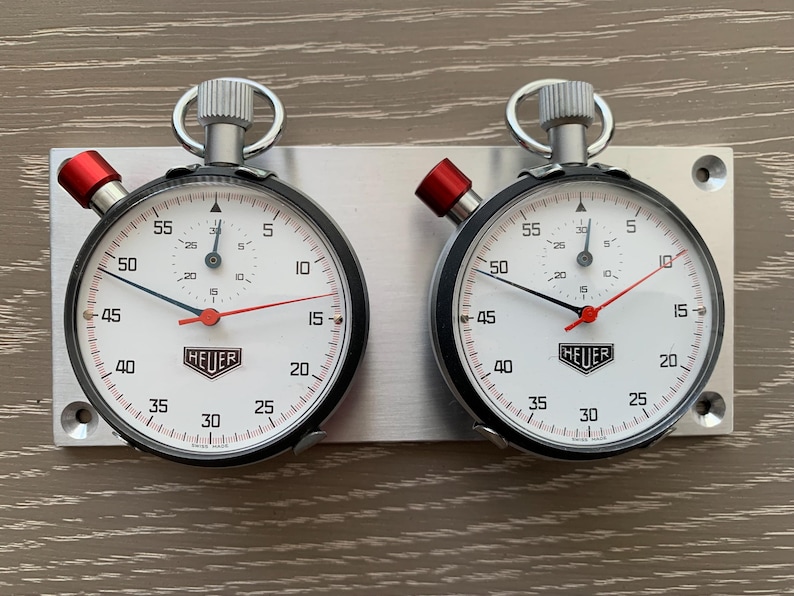 May include: Two vintage Heuer stopwatches mounted on a silver plate. Each stopwatch features a white face with black numerals, a red second hand, and a black bezel. The stopwatches are designed for precision timing.