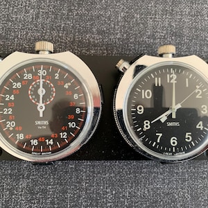 Może przedstawiać: Dwa zabytkowe stopery Smiths. Jeden ma czarno-czerwoną tarczę, drugi czarną tarczę z białymi cyframi. Oba są w srebrnych obudowach i zamontowane na czarnej podstawie. Widoczny napis 'Great Britain'.