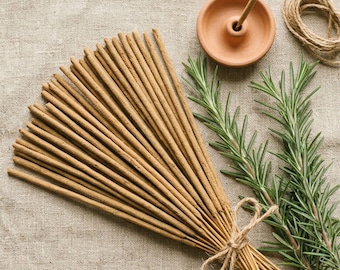 May include: A bundle of light brown incense sticks tied with twine, accompanied by a small terracotta incense holder with a single stick inserted, and fresh green rosemary sprigs. The items are arranged together on a textured fabric surface.