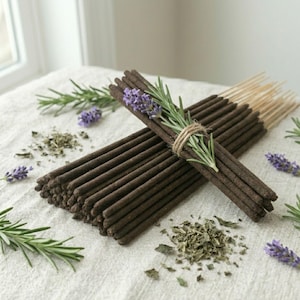 May include: Bundles of brown incense sticks are arranged on a white cloth, accented with sprigs of rosemary, lavender buds, and dried herbs. One bundle is tied with twine and adorned with fresh lavender and rosemary. The incense sticks are approximately 10 inches long.
