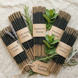 May include: Bundles of incense sticks with black bodies and tan tips are arranged with sprigs of herbs. Each bundle is labeled with a kraft paper band or tag. The labels read: Lavender Calm, Clary Sage Clarity, Peppermint Revive, Rosemary Restore, Sage & Lavender Unwind, and Carolina Rooted.