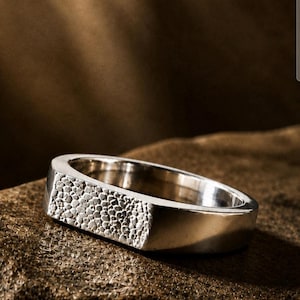 May include: A silver ring with a textured rectangular top. The ring is set against a brown, textured background, highlighting its polished metal finish. The ring's design is simple yet striking.