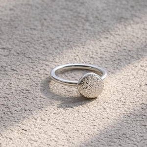 May include: A silver ring with a textured, circular design. The ring band is smooth and thin, leading to a raised, textured circle. The ring is set against a neutral, textured background.