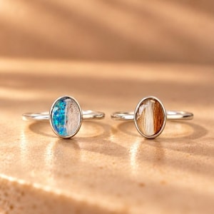 May include: Two silver rings featuring oval settings. The left ring contains a split inlay of iridescent blue flecks and light-colored fibers. The right ring features an inlay of brown and white fibers encased in a smooth, clear material.