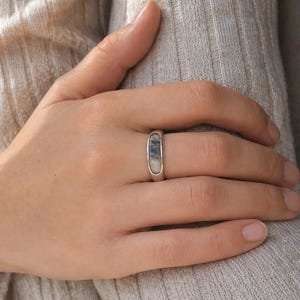May include: A silver ring with a rectangular, stone-like inlay. The inlay features a mix of grey and white tones. The ring is shown on a hand, with a neutral-toned background.