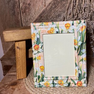 Puede incluir: Marco de fotos rectangular con un diseño floral de narcisos. El marco presenta un fondo rosa claro con narcisos blancos y amarillos y hojas verdes. El marco está sobre un salvamanteles tejido.