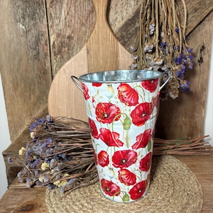 Könnte beinhalten: Ein Metalleimer mit einem floralen Mohnblumenmuster. Der Eimer hat einen weißen Hintergrund mit leuchtend roten Mohnblumen und grünem Laub. Er hat zwei kleine Metallgriffe und steht auf einer gewebten Matte. Trockenblumen und ein Holzbrett befinden sich im Hintergrund.