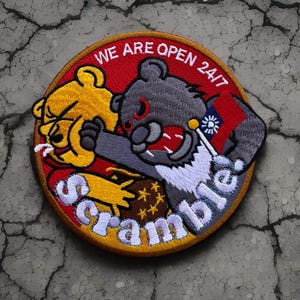 May include: A round embroidered patch with a red background and the text "WE ARE OPEN 24/7". A cartoon bear is being attacked by a cartoon raccoon. The raccoon is holding a flag with a white sun and a blue background. The text "Scramble!" is embroidered in white on a yellow background.
