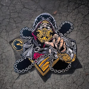 May include: A black and yellow embroidered patch featuring a hooded figure with a yellow mask and chains. The figure is holding out a hand with a fist clenched.
