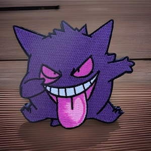 May include: Embroidered patch featuring a purple cartoon ghost with a wide grin, pink eyes, and a long pink tongue. The patch has a detailed stitched design and is likely for application on clothing or accessories.