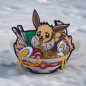 May include: Embroidered patch featuring a cartoon Eevee character enjoying ramen. The patch includes a bowl of ramen with noodles, eggs, and other ingredients. The Eevee character is brown, tan, and white, with a cute expression. The patch is set against a snowy background.
