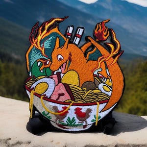 May include: An embroidered patch featuring a cartoon orange and red fire-breathing dragon in a bowl of ramen noodles. The dragon is surrounded by various toppings, including eggs, meat, and vegetables. The bowl is decorated with a white and green pattern.