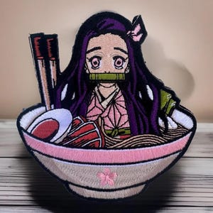 May include: Embroidered patch featuring a cartoon anime character with purple hair, wearing a pink and purple kimono, sitting in a bowl of ramen noodles. The character has a green mouth guard and is holding chopsticks. The bowl is decorated with a pink rim and a pink cherry blossom.