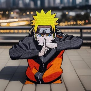 May include: An embroidered patch featuring a cartoon character with yellow hair, blue eyes, and an orange and black outfit. The character is making a hand gesture with their index finger to their lips, suggesting silence.