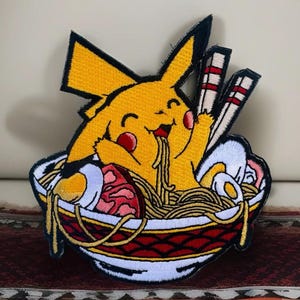 May include: A yellow Pikachu cartoon character is eating ramen noodles from a red and white bowl. The bowl is filled with noodles, eggs, and meat. There are chopsticks on the side of the bowl.