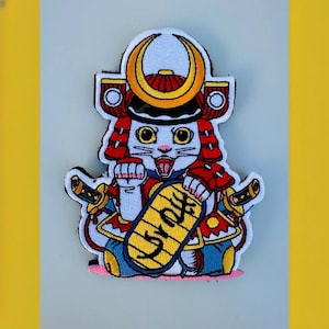May include: Embroidered patch of a white cat wearing a red and gold samurai helmet, holding a yellow sign with Japanese characters. The cat has a fierce expression and is holding two swords.