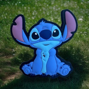 May include: A blue cartoon character with large ears, big eyes, and a friendly expression. The character is sitting on a patch of green grass. The character has a black outline and pink inner ears.