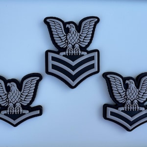 May include: Three black and gray military patches featuring a detailed eagle emblem atop a chevron design. The patches are arranged on a white background, showcasing the intricate details of the eagle and chevron.