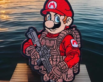 Puede incluir: Parche bordado que representa a un personaje de dibujos animados con gorro y uniforme rojos, sosteniendo un rifle. El personaje tiene bigote y lleva un chaleco táctico. El parche está sobre un fondo de agua y un muelle de madera.