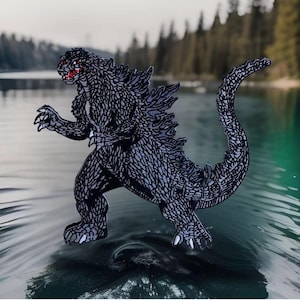 May include: A black and grey embroidered patch of Godzilla, a fictional monster, standing on a lake with a forest in the background.